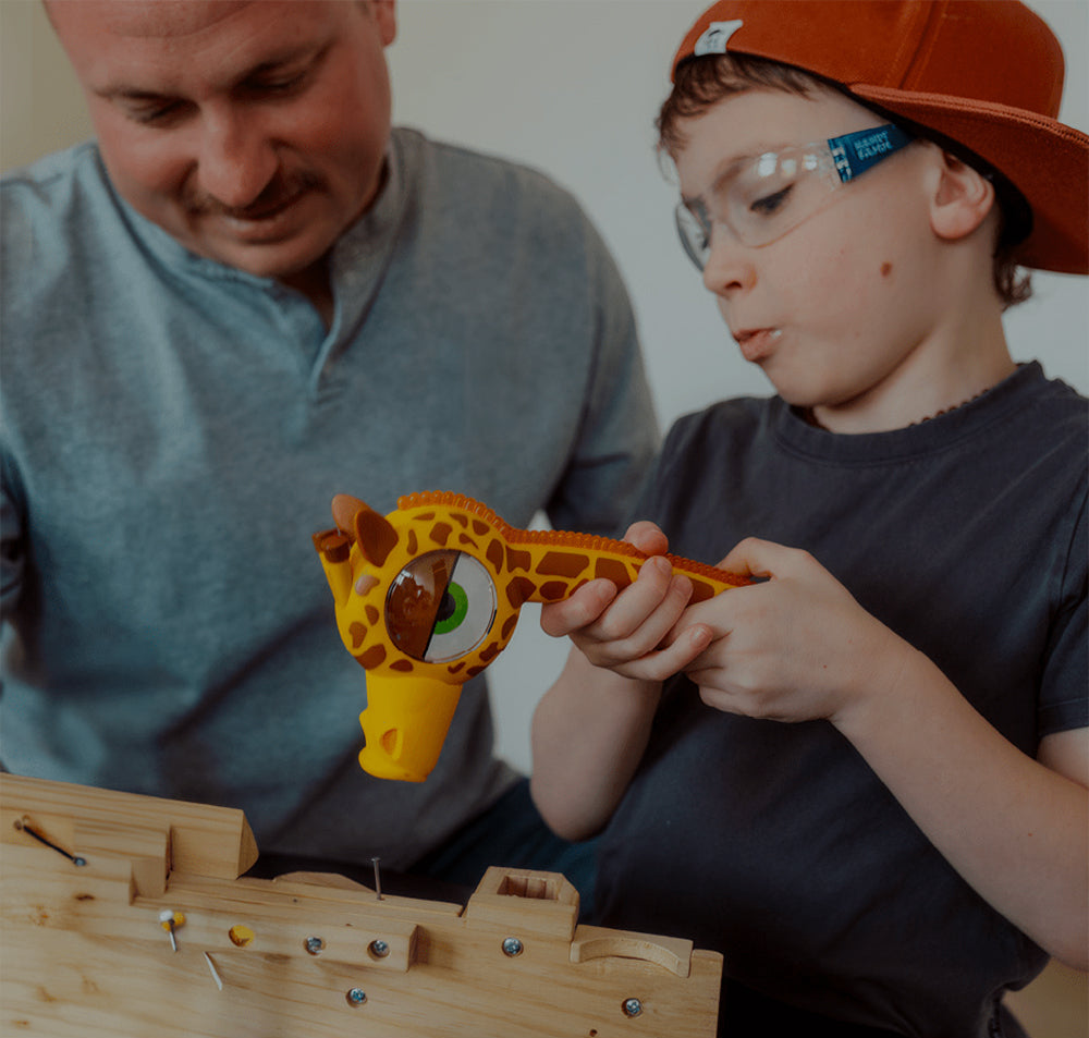 A young boy wearing a brown cap and safety glasses holds a giraffe-shaped toy microscope over a wooden model while a man watches closely.